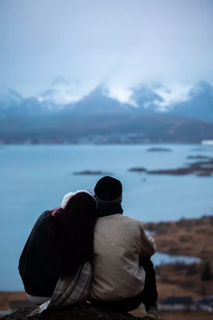 Couple sitting closely on a mountaintop, admiring fjord views in Lofoten under winter skies.
