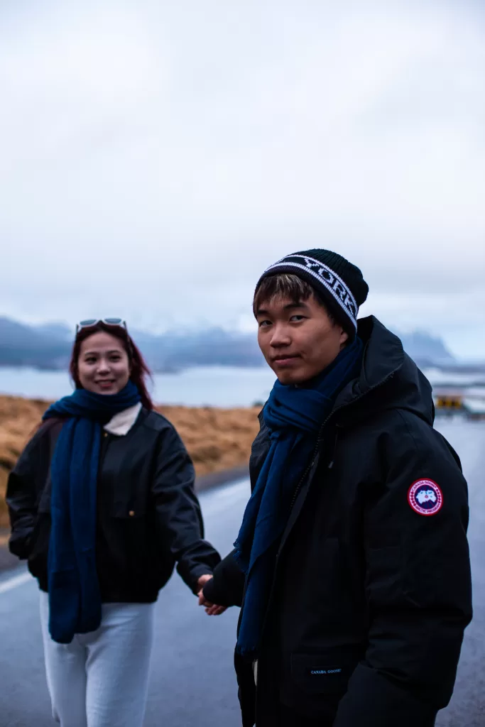 Couple holding hands on a scenic Arctic road in Lofoten, dressed in cozy winter jackets.