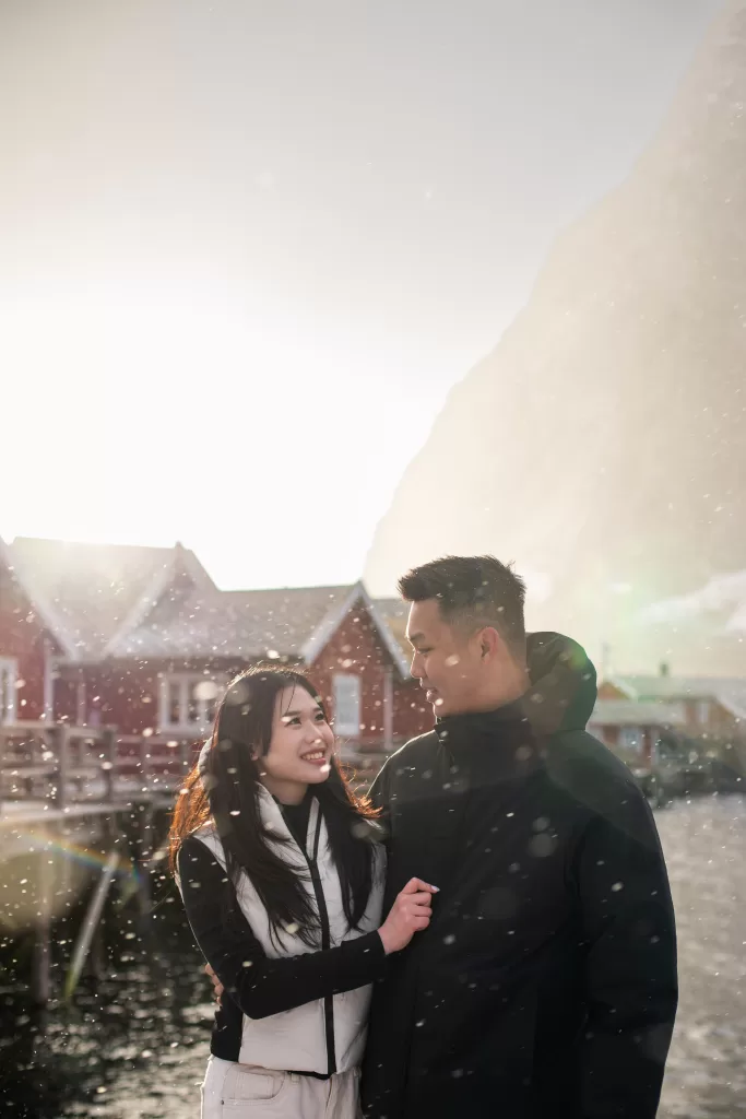 Couple smiling during snowfall in Lofoten fishing village with red rorbuer cabins in the background.