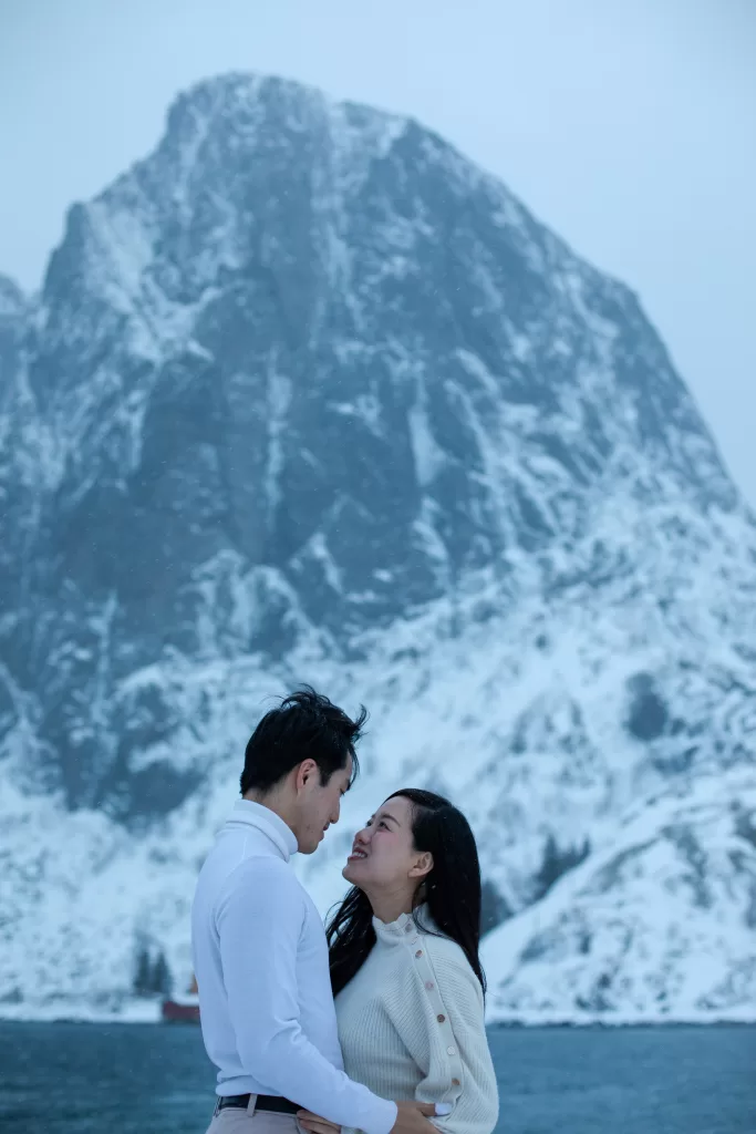 Romantic couple looking at each other in front of a dramatic snowy mountain in Lofoten during a winter photoshoot.