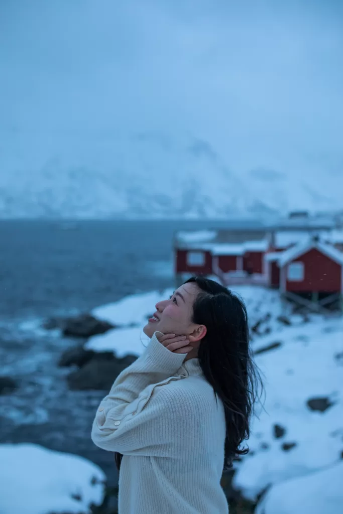 Smiling woman enjoying snowfall in Hamnøy, Lofoten, with traditional red fishing cabins and snowy coastline in the background.
