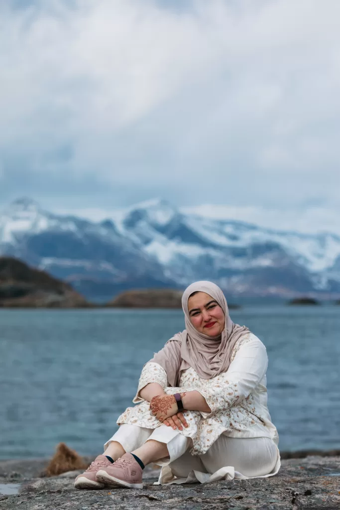 Smiling woman in a hijab seated on rocks by the water with snow-capped mountains near Henningsvær, Lofoten.