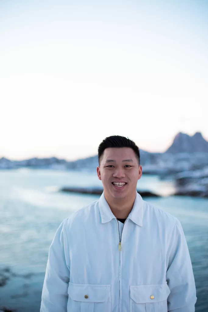 Man smiling in soft Arctic light with scenic snowy fjord and Lofoten mountain silhouettes in the background.