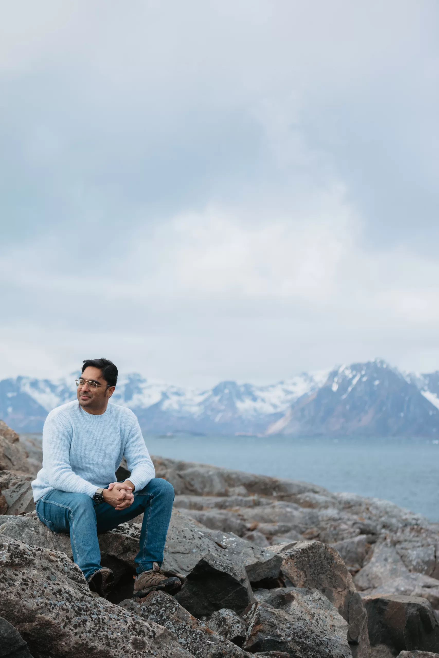 Man sitting on coastal rocks in Northern Norway with scenic snowy mountains and Arctic fjord in the background.