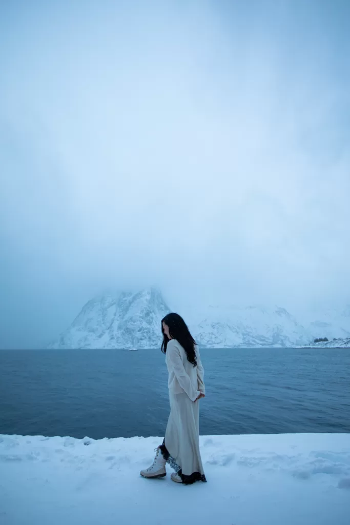 Woman in winter outfit walking along a snowy shore with mist-covered mountains and Arctic fjord in Lofoten, Norway.