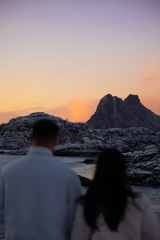 Silhouetted couple gazing at snow-dusted rocks and mountains under pastel Arctic sky in Lofoten during a peaceful sunset.