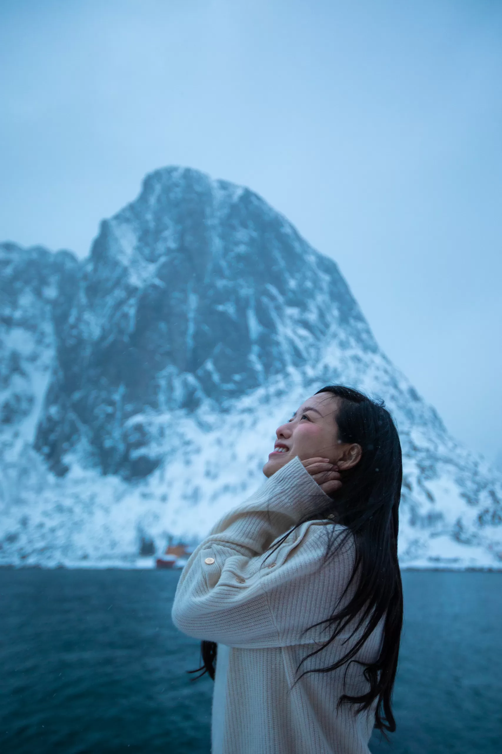 Portrait of a woman smiling under snowfall with towering snow-covered cliffs behind her in Lofoten, Arctic Norway.