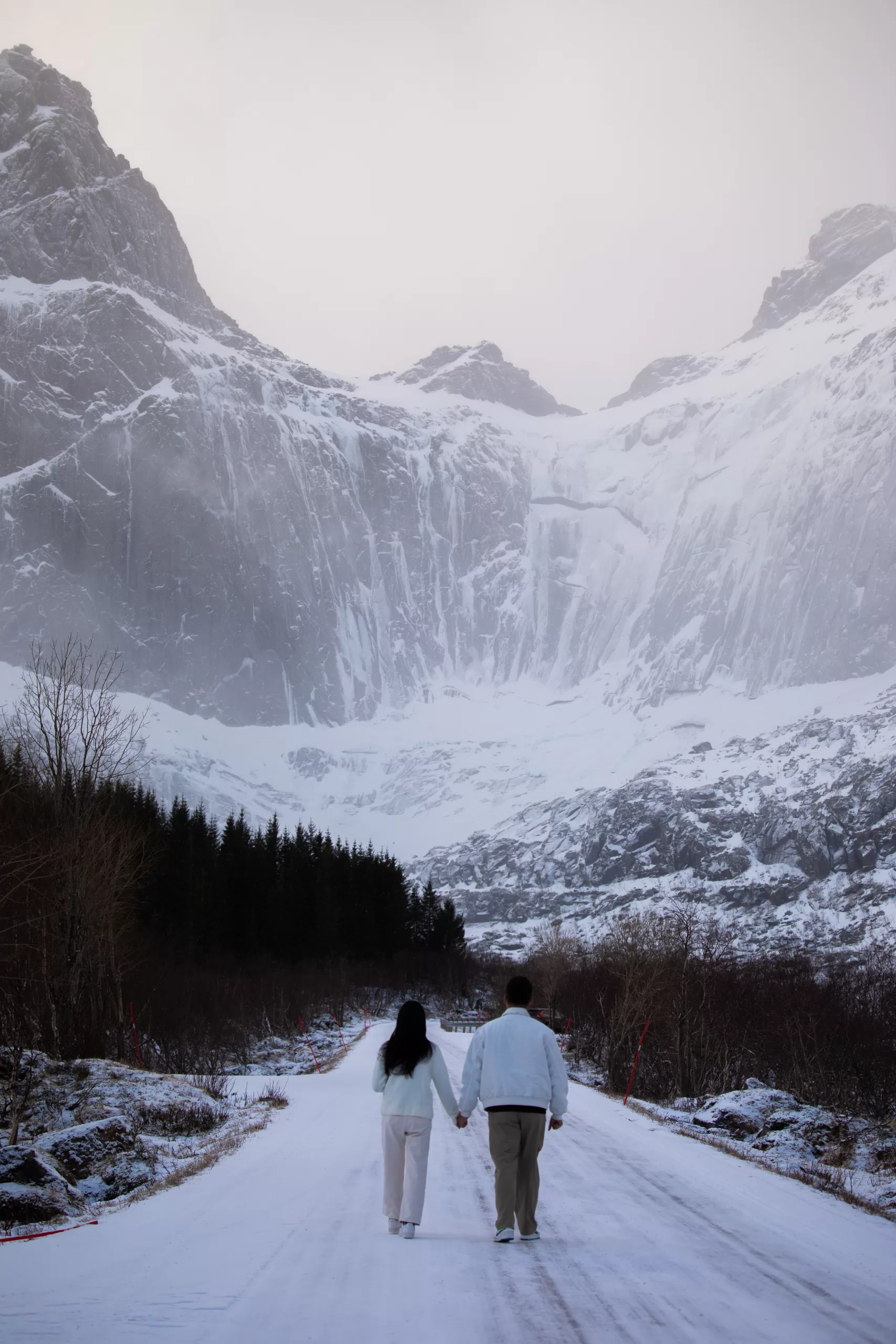 Couple walking along a snow-covered path in Lofoten with dramatic mountain cliffs in the background.